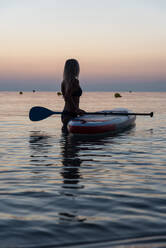 Seitenansicht einer nicht erkennbaren weiblichen Surferin, die in der Nähe des Paddelboards steht und gegen die spektakuläre Sonne im Sonnenuntergang rudert - ADSF16345