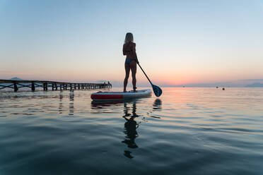 Back view of silhouette of unrecognizable female surfer standing on paddleboard and rowing against spectacular sun in sunset sky - ADSF16348