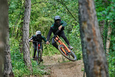 Männliche Radfahrer in Schutzhelmen und Kostümen, die gefährliche Stunts auf Fahrrädern beim Downhill auf einem Waldweg ausführen - ADSF16362