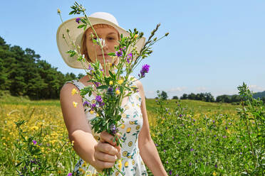 Zufriedene Frau in Sommerkleid und Hut steht mit einem Strauß Wildblumen auf einer blühenden Wiese und schaut in die Kamera - ADSF16365