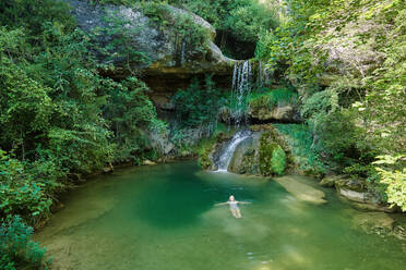 From above female traveler in bikini lying in lake near waterfall in forest and enjoying summer holiday - ADSF16367