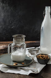 Glas und Flasche mit veganer Milch und Schale mit Mandeln stehen auf dem rustikalen Tisch neben Serviette und Sieb - ADSF16392