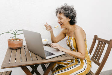 Lächelnde reife Frau, die am Tisch an einer weißen Wand im Hinterhof isst und dabei einen Laptop benutzt - TCEF01196