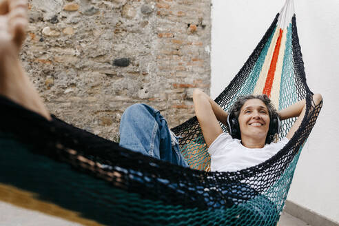 Smiling woman listening music through headphones while relaxing on hammock against wall at back yard - TCEF01199