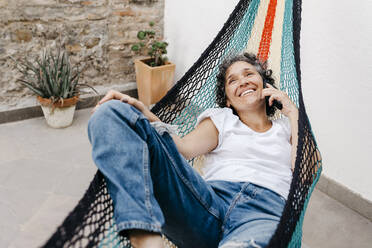 Lächelnde Frau, die über ihr Smartphone spricht, während sie sich in einer Hängematte im Garten entspannt - TCEF01201
