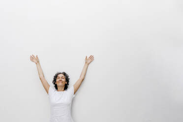 Lächelnde reife Frau steht mit geschlossenen Augen und erhobenen Armen vor einer weißen Wand - TCEF01205