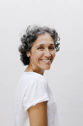 Happy mature woman with short curly hair standing against white wall - TCEF01206