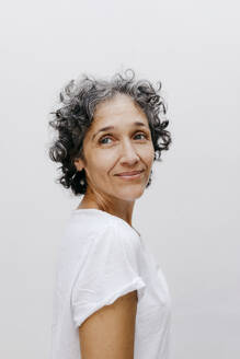 Mature woman with short curly hair looking away while standing against white wall - TCEF01207