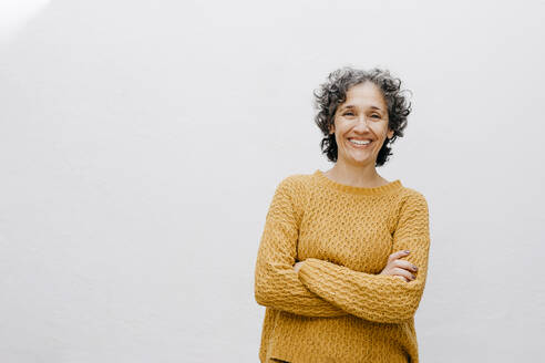 Smiling woman in orange sweater standing with arms crossed against white wall - TCEF01209