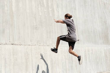 Seitenansicht eines mutigen jungen Mannes, der auf die Betonwand eines Gebäudes springt, während er einen gefährlichen Stunt macht und Parkour in der Stadt betreibt - ADSF16420