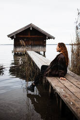 Weibliche Reisende in warmer Kleidung sitzt auf Holzplanke Pier in der Nähe von schäbigen Schuppen befindet sich am See in düsteren Herbsttag - ADSF16423