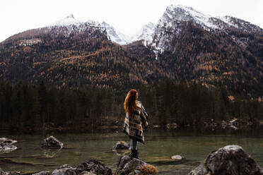 Side view of unrecognizable distant female traveler in warm outfit standing on rocky shore of lake and observing amazing scenery of autumn forest and snowy mountains in cloudy day - ADSF16425