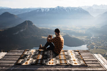 Rückenansicht eines nicht erkennbaren männlichen Touristen, der auf einer Decke am Aussichtspunkt sitzt und ein Foto macht - ADSF16430