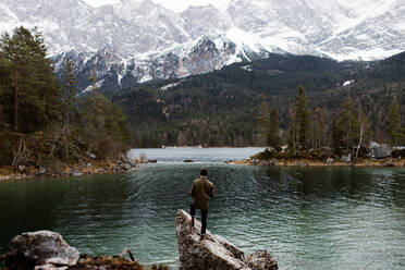 Back view of man standing on stone on shore of pond and observing mountains covered with snow in winter - ADSF16445