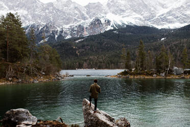 Rückenansicht eines Mannes, der auf einem Stein am Ufer eines Teiches steht und die schneebedeckten Berge im Winter betrachtet - ADSF16445