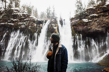 Mann stehend auf malerische Landschaft von mächtigen Wasserfall mit Pool fließt unter verschneiten Wald in gebirgigem Gelände im Winter Tag in den USA Blick weg - ADSF16454