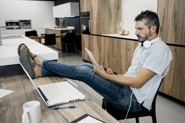 Älterer männlicher Freiberufler, der ein digitales Tablet hält, während er mit hochgelegten Füßen auf einem Schreibtisch im Büro sitzt - RCPF00292