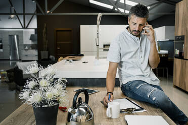 Gutaussehender männlicher Fachmann, der über sein Mobiltelefon spricht, während er mit einem Klemmbrett auf dem Schreibtisch im Büro sitzt - RCPF00294