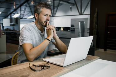 Thoughtful mature businessman working while looking at laptop in office - RCPF00296