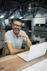 Cheerful handsome male freelancer sitting with laptop at desk while working in office - RCPF00297