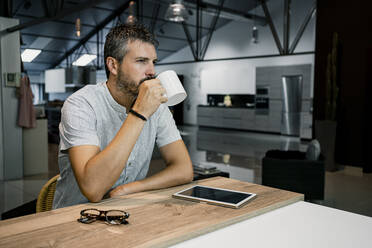 Thoughtful male entrepreneur drinking coffee while sitting with digital tablet and eyeglasses at desk in office - RCPF00299