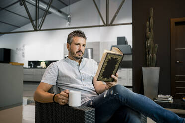 Entspannter männlicher Freiberufler, der mit einer Kaffeetasse auf einem Stuhl im Büro sitzt und ein Buch liest - RCPF00305