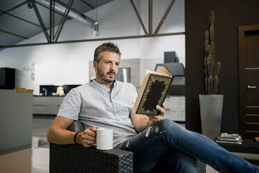 Relaxed male freelancer reading book while sitting with coffee cup on chair in office - RCPF00305