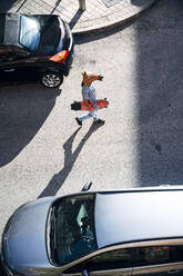 Mittlerer erwachsener Mann mit Pferdemaske, der ein Skateboard hält, während er inmitten von Autos auf einer Straße in der Stadt spazieren geht - JCMF01534