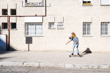 Mann mit Pferdemaske beim Skateboardfahren auf dem Fußweg vor einem Gebäude in der Stadt an einem sonnigen Tag - JCMF01536