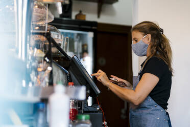 Inhaberin mit Gesichtsschutzmaske an der Kasse eines Cafés - EGAF00853