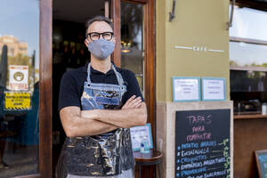 Male owner in face mask standing with arms crossed against cafe during COVID-19 crisis - EGAF00854