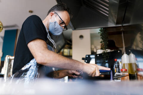 Männlicher Barista mit Gesichtsmaske beim Kochen in der Küche eines Cafés während der COVID-19-Krise, lizenzfreies Stockfoto