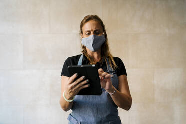 Weiblicher Besitzer mit Gesichtsschutzmaske, der ein digitales Tablet an der Wand eines Cafés benutzt - EGAF00869