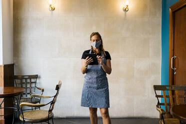 Weibliche Café-Besitzerin trägt einen Gesichtsschutz, während sie ein digitales Tablet an der Wand benutzt - EGAF00870