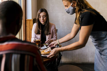 Kellnerin mit Gesichtsschutz, die einem jungen Paar in einem Café während COVID-19 das Essen serviert - EGAF00883