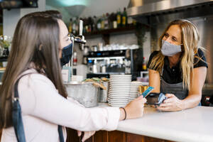 Businesswoman in face mask paying through credit card at counter in cafe during COVID-19 crisis - EGAF00898