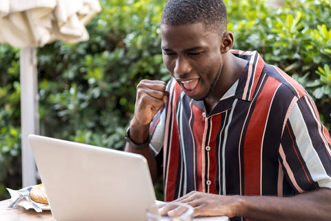 Happy young man clenching fist while using laptop on terrace of cafe stock photo