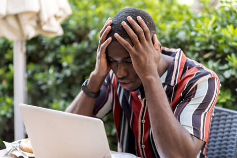 Tensed young man sitting with head in hands while looking at laptop in cafe stock photo