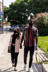 Young couple in face masks holding hands while walking in city during COVID-19 crisis - EGAF00926