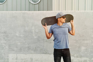 Entschlossener männlicher Skater in trendigem Outfit und Sonnenbrille steht mit Skateboard auf den Schultern in der Stadt und schaut weg - ADSF16501