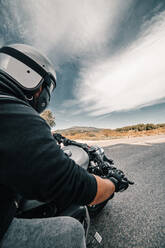 Back view cropped unrecognizable male rider in leather jacket and helmet riding motorbike on roadside against cloudy sky in countryside - ADSF16560