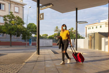 Junge Frau mit Schutzmaske beim Gehen mit Gepäck an der Bushaltestelle - JPTF00587