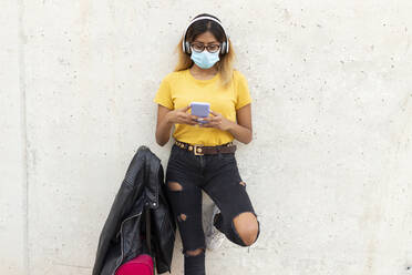 Junge Frau mit Schutzmaske bei der Benutzung eines Smartphones mit Gepäck an einer weißen Wand an einer Bushaltestelle - JPTF00588