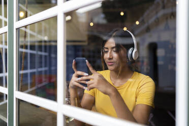 Schöne junge Frau hört Musik und benutzt ihr Smartphone in einer Bar, gesehen durch ein Fenster - JPTF00591