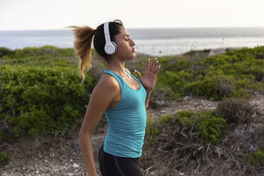 Junge lateinamerikanische Frau hört Musik beim Joggen am Meer - JPTF00595