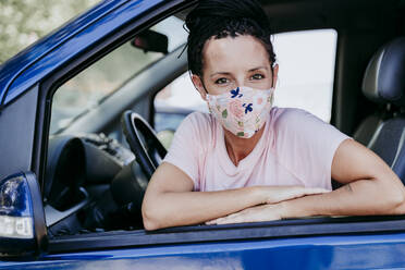 Woman wearing floral protective mask sitting in car while leaning on window - EBBF00792