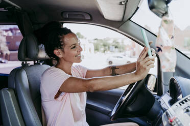 Lächelnde Frau, die ein Selfie macht, während sie an einem sonnigen Tag im Auto sitzt - EBBF00793