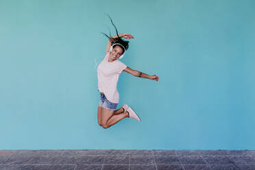 Fröhliche Frau springt beim Musikhören über Kopfhörer gegen eine türkisfarbene Wand - EBBF00803
