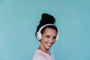 Lächelnde schöne Frau hört Musik über Kopfhörer vor türkisem Hintergrund - EBBF00807