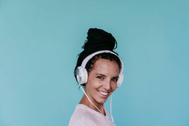 Smiling beautiful woman listening music through headphones against turquoise background - EBBF00807